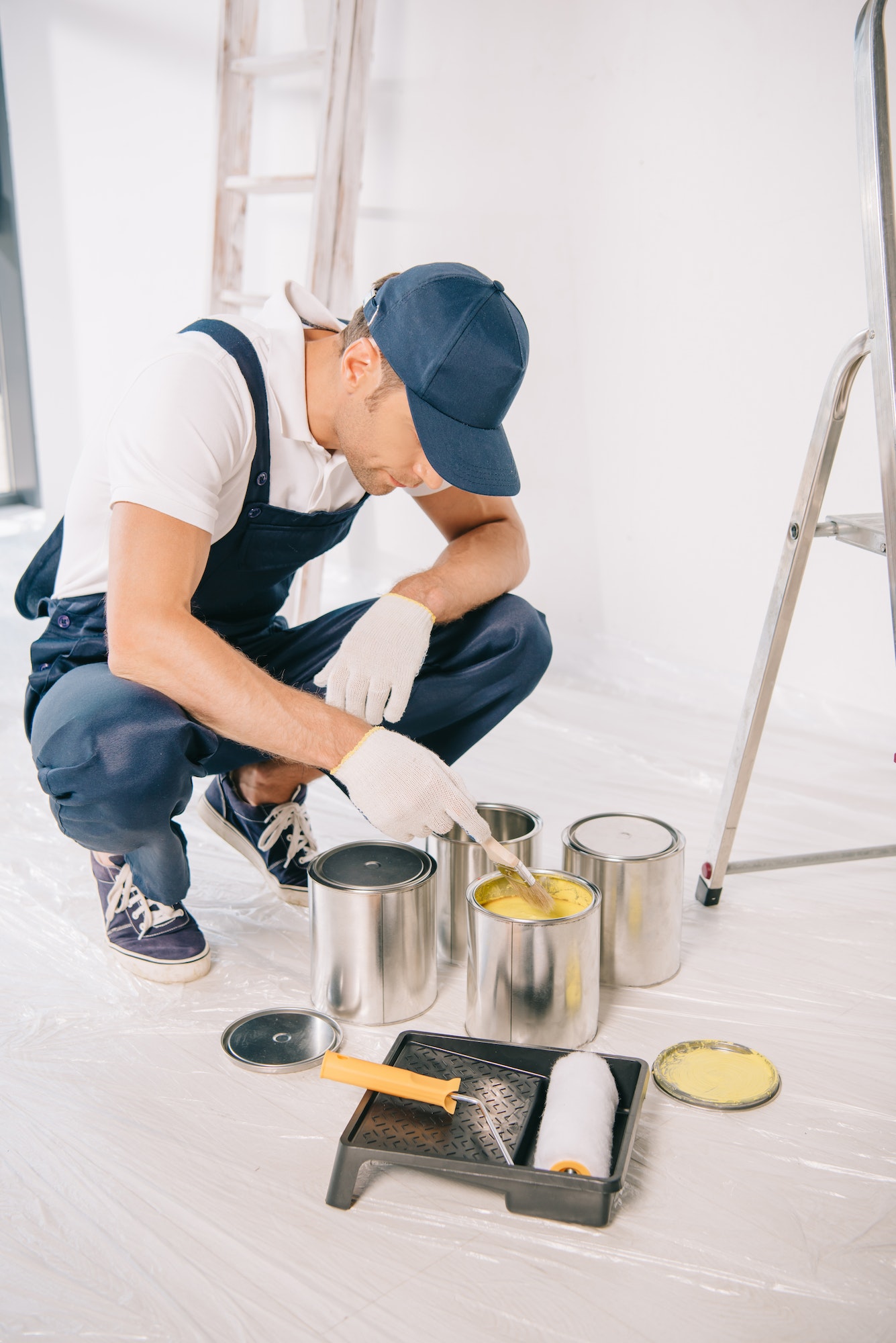 young painter in overalls and cap taking yellow paint from can with paintbrush